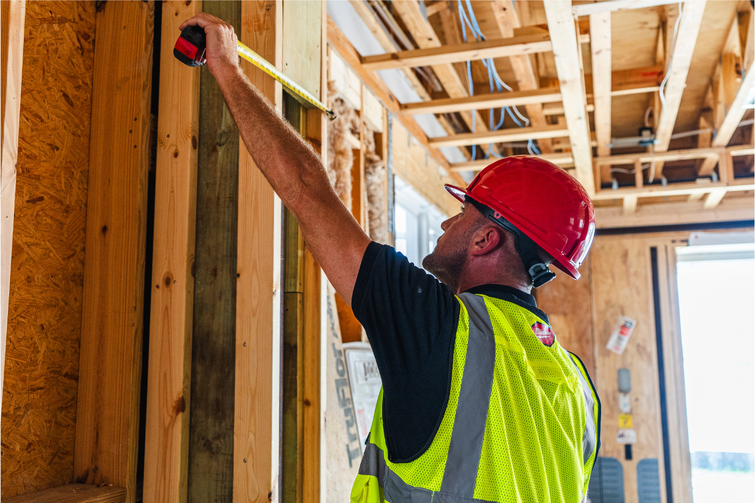 Construction worker in a red hard hat and high-visibility vest takes a wall measurement with a tape measure inside a framed building.