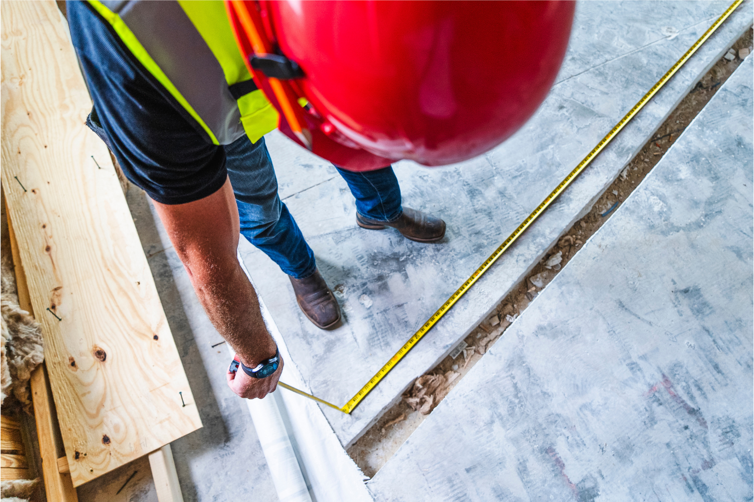 Top-down view of a construction worker in a safety vest and red hard hat measuring a concrete floor with a tape measure.