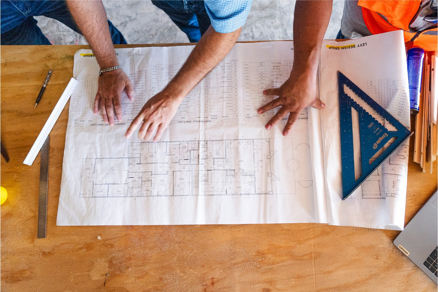 Hands spread over architectural blueprints on a wooden table, with rulers and a blue triangular ruler nearby.
