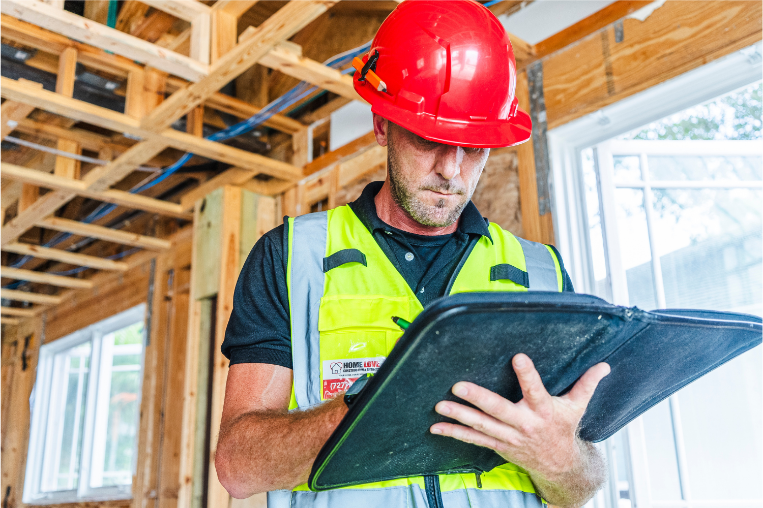 Construction worker in a high-visibility vest and red hard hat reviews blueprints on a clipboard inside an unfinished wooden room.