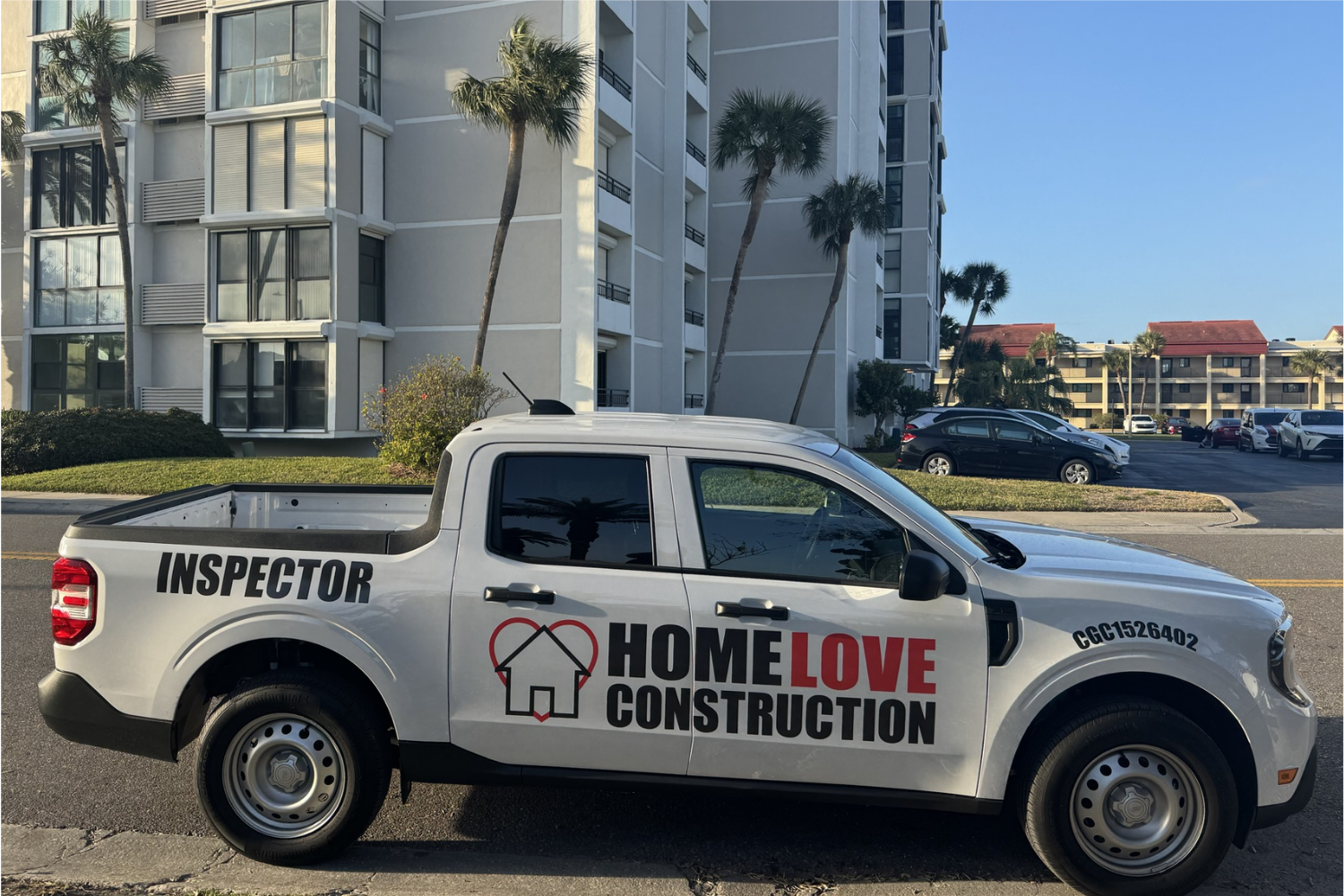 White pickup truck with 'INSpector' on the rear and 'HOME LOVE CONSTRUCTION' logo parked on a street in front of a modern, light-gray apartment building with tall palm trees nearby.