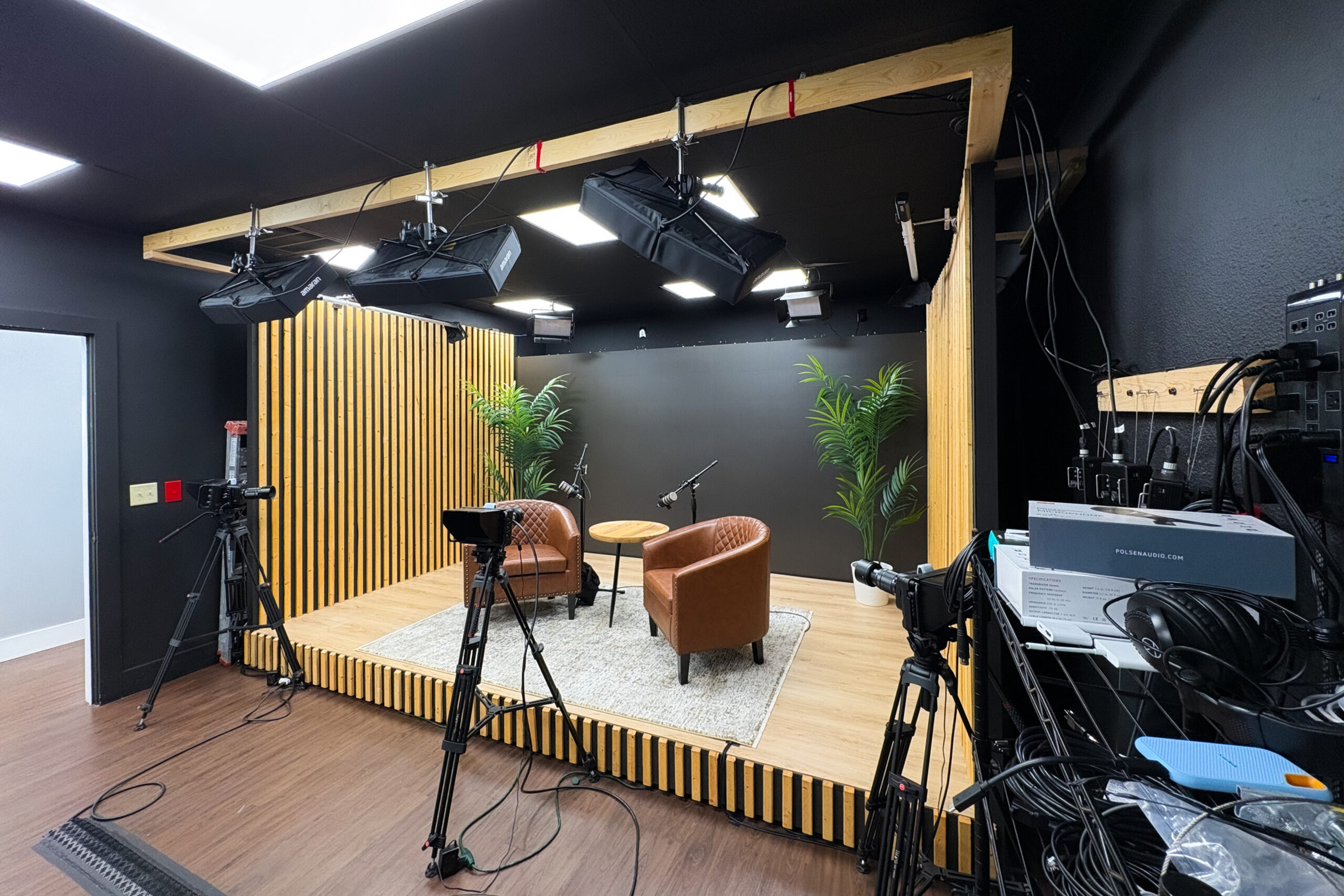 Studio set with stage, chairs, and camera rigs facing a black wall and wooden slat backdrop